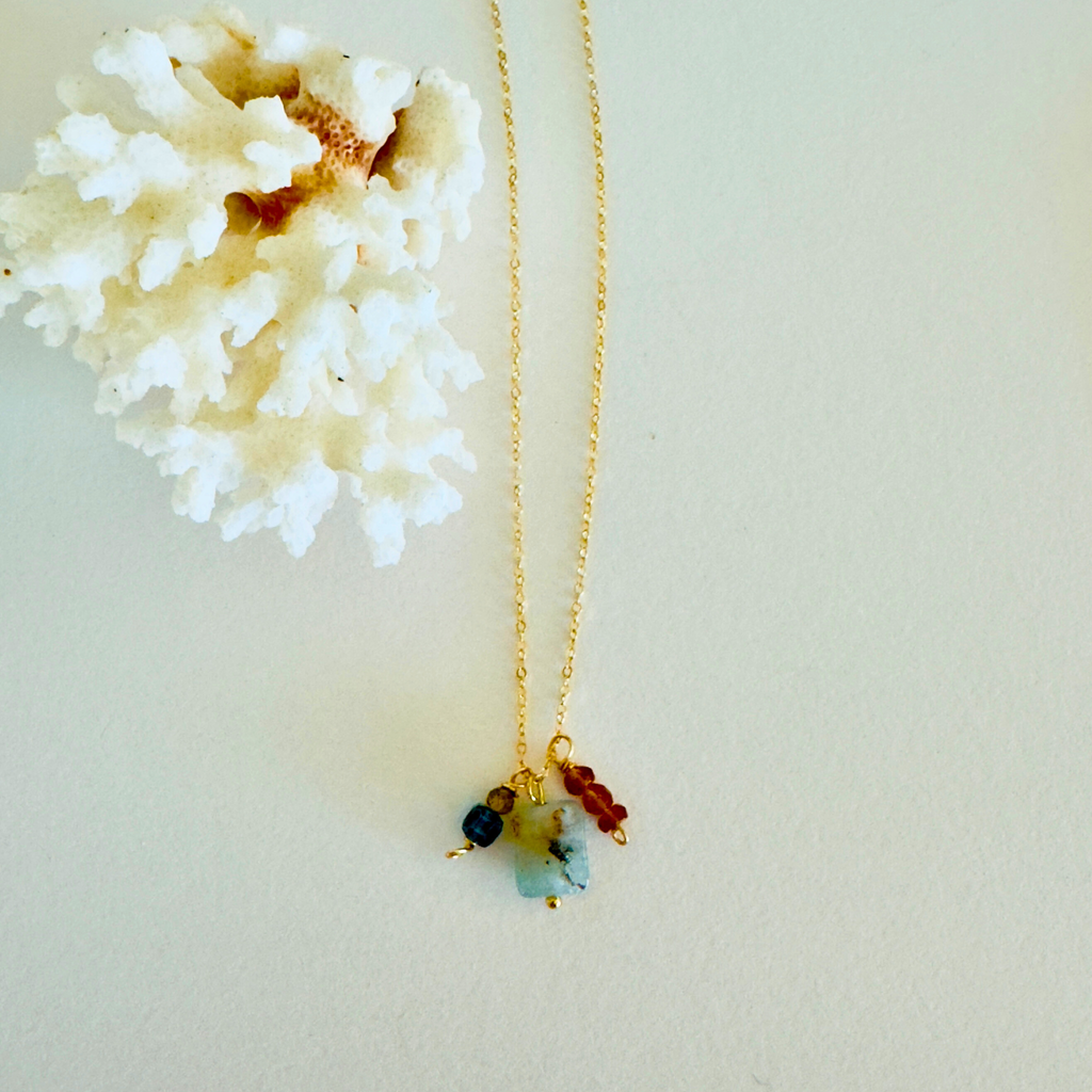 A necklace is displayed on a cream white background and framed by white coral. Genuine turquoise, aventurine, garnet hessonite, and mystic topaz stones form a pendant at the end of a dainty 14k gold filled chain.